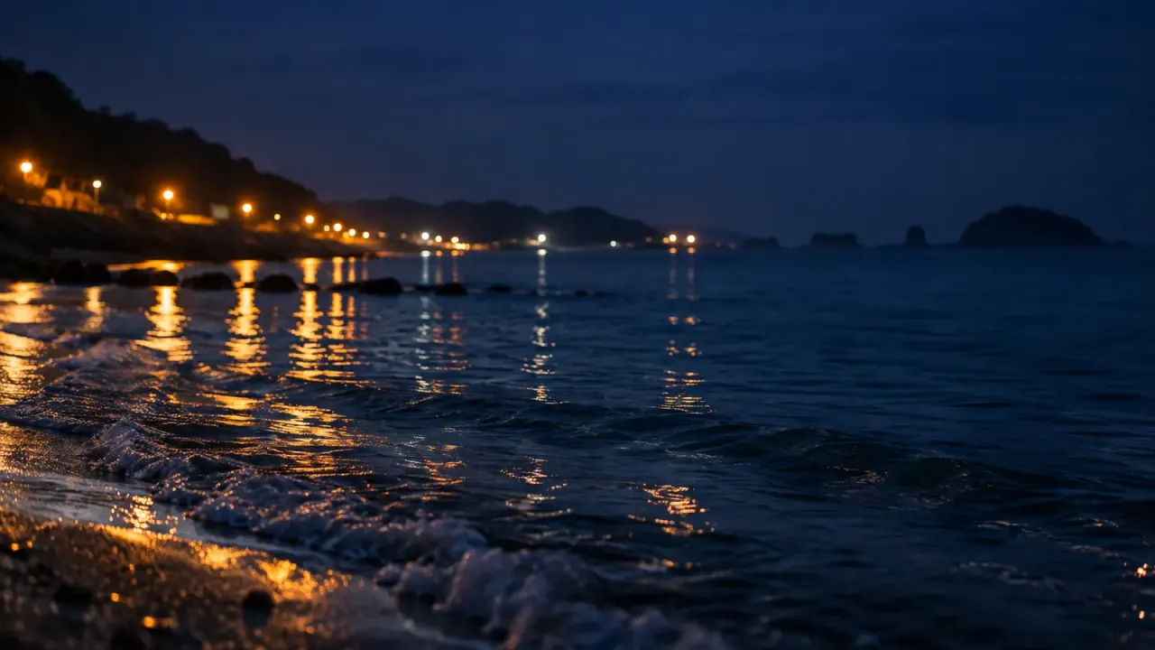 夜の海辺に、岸に沿って橙色の灯りが並び、水面に映る(イメージ)